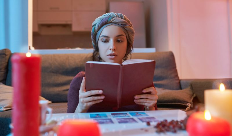 Serious beautiful Caucasian fortune-teller with a book in her hands sitting at the divination table